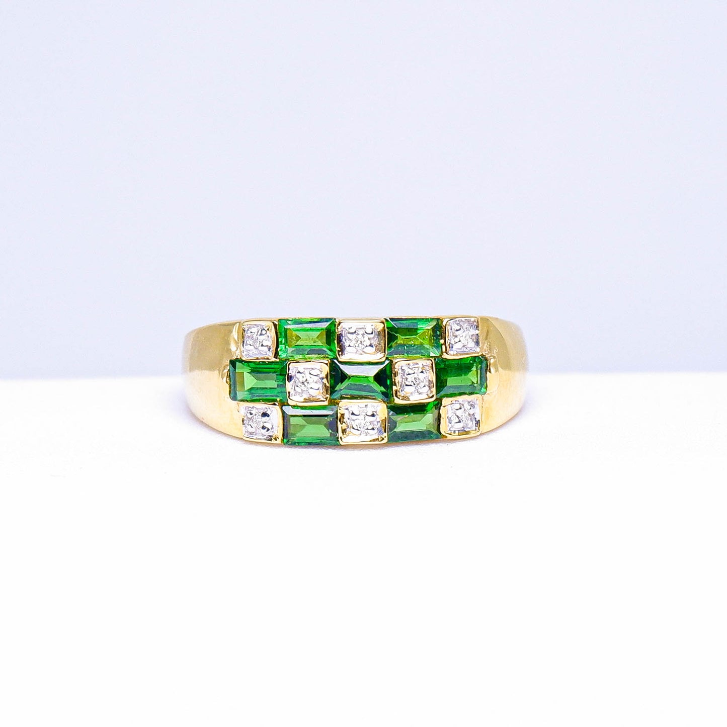 Gold ring with square green gemstones and white stones in a grid pattern on white background